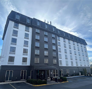 A tall modern multi-story hotel or apartment building with white and dark gray exterior walls, many windows, a dark roofline, and a paved parking area in front under a blue sky with clouds.