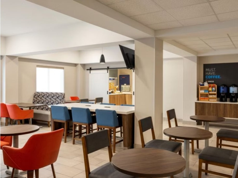 A modern cafe or breakroom interior with round wooden tables, blue and orange chairs, a counter, and a coffee station in the background.