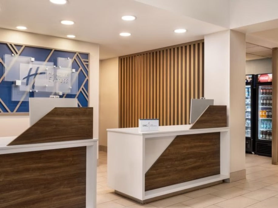 A modern hotel lobby reception desk with white surfaces and dark wood paneling, set in a bright interior with tiled floors and a slatted wood accent wall behind it.