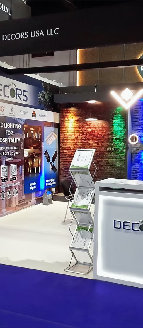A booth display for Decors USA LLC at an exhibition, featuring a white reception desk, a freestanding brochure stand, LED lighting samples, and a brick wall backdrop with colored lights.