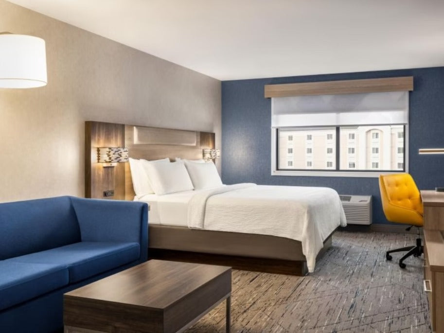 A modern hotel room featuring a bed with white linens, a blue sofa, a blue accent wall, a yellow office chair, and a large window looking out onto another building.