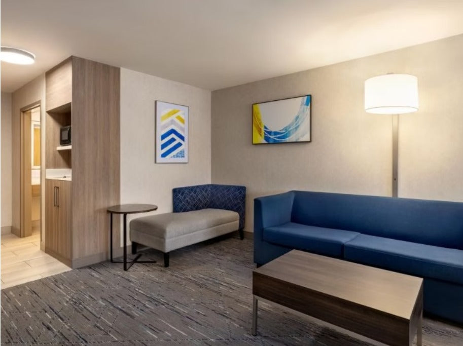 A modern hotel suite living room featuring a blue sofa, a matching blue accent chair, a gray bench, a wooden coffee table, and framed abstract wall art.