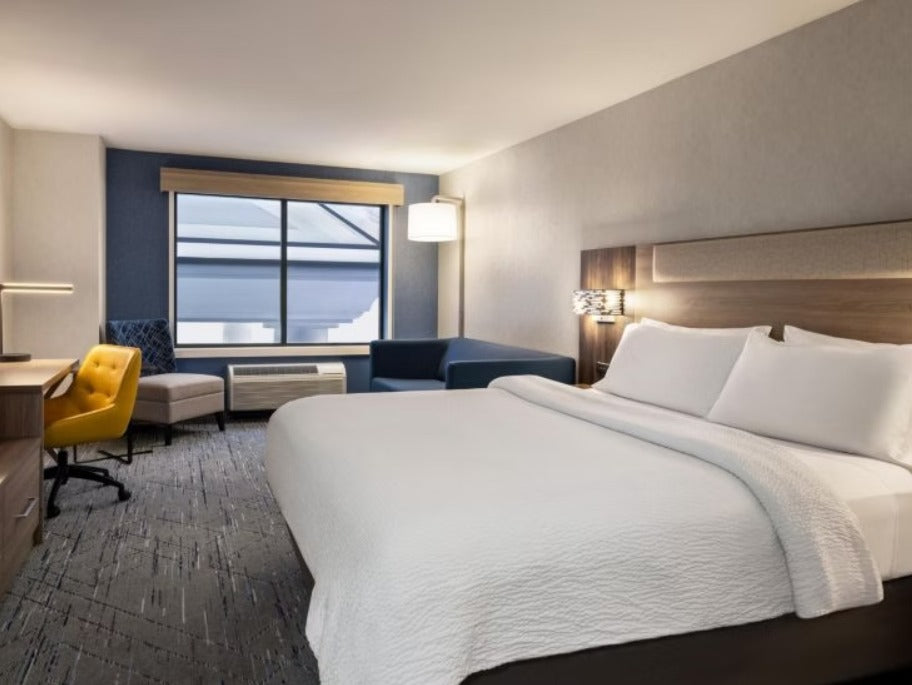 A modern hotel room featuring a large bed with crisp white bedding, a dark wood headboard, a gray carpet, a blue accent wall, and a yellow chair near a window.