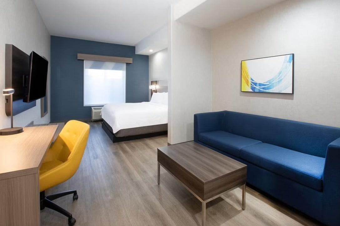 A modern hotel suite living area featuring a blue sofa, a wooden coffee table, a yellow office chair, a desk with a mounted TV, and a bed in the background.