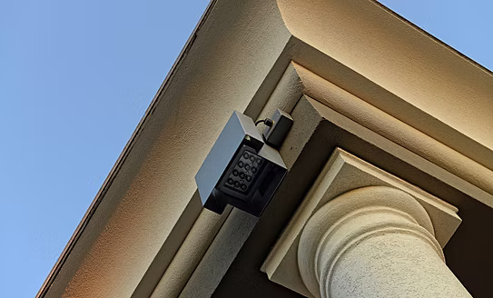 A black electronic keypad security camera mounted on the corner of a beige stucco building column against a clear blue sky.