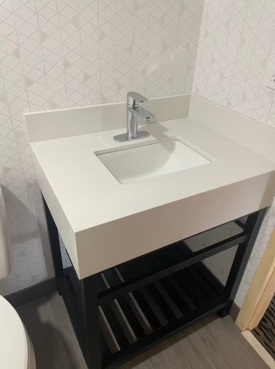 A modern white vanity sink with a rectangular basin and a chrome faucet, mounted on a black metal frame with a lower shelf, set against a light patterned tiled wall.