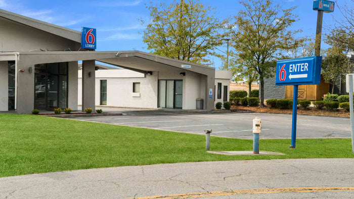 A Motel 6 entrance with a modern single-story building, grassy lawn, parking lot, and blue sign with the Motel 6 logo and an 'ENTER' arrow.