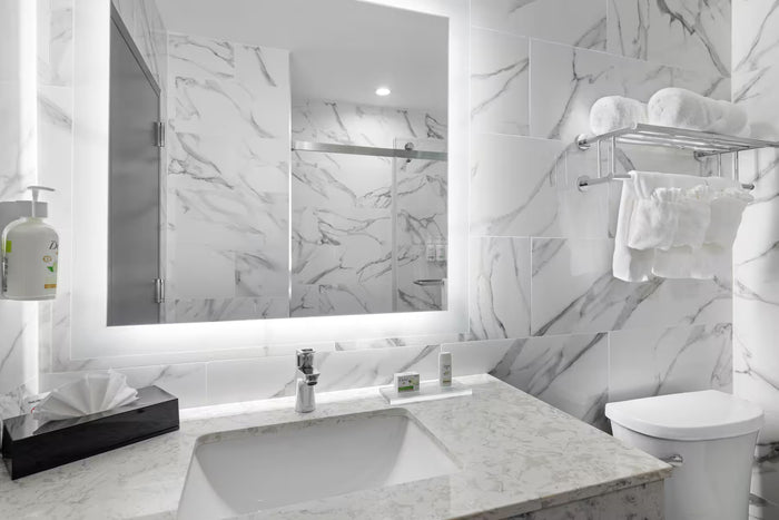 A modern bathroom vanity with a white marble countertop and matching marble wall tiles, featuring a large backlit mirror, chrome faucet, toilet, and towel rack with white towels.