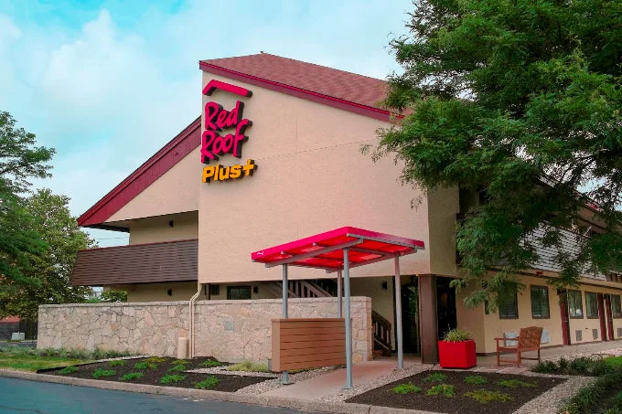 Red Hat Plus+ restaurant building exterior with a beige facade, red roof, large red and yellow logo, stone wall, covered entrance, and surrounding trees.