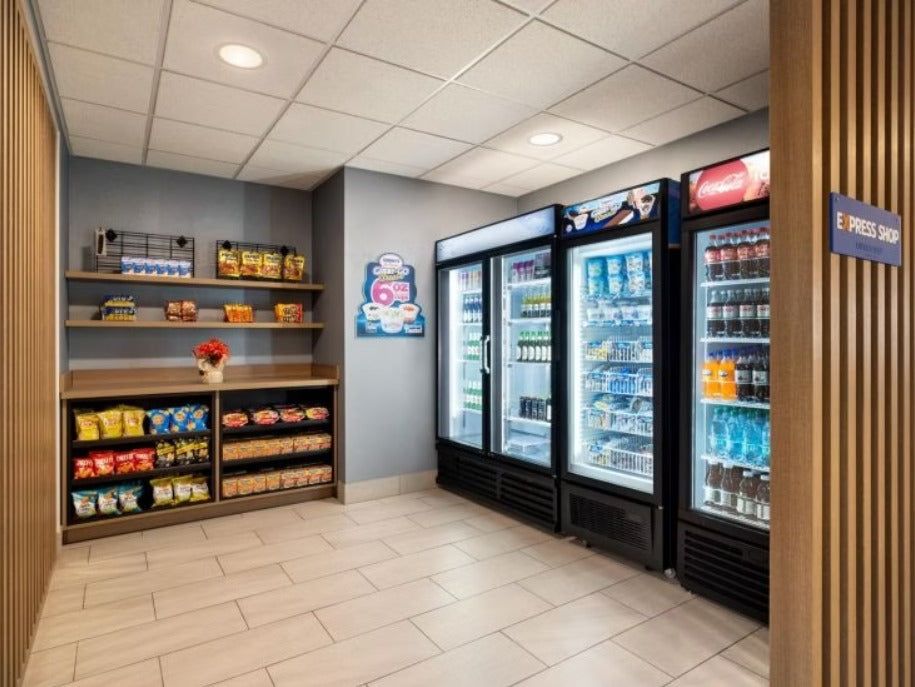 A clean, modern convenience store or breakroom area with three glass-door beverage refrigerators stocked with bottled drinks, a small snack and candy display shelf on the left, tiled floor, and wood-paneled walls.