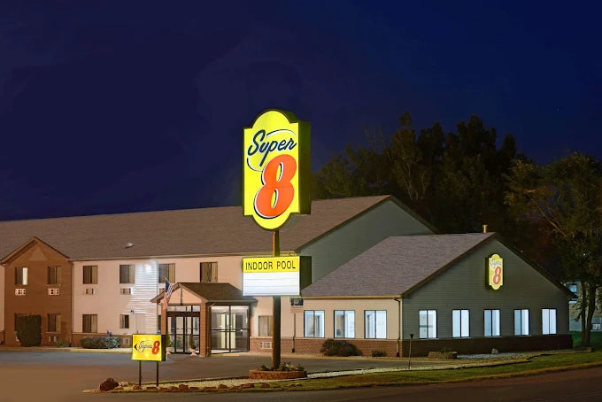 Super 8 motel building at night with a large illuminated Super 8 sign on a pole, showing the front entrance, parking area, and surrounding trees under a dark blue sky.
