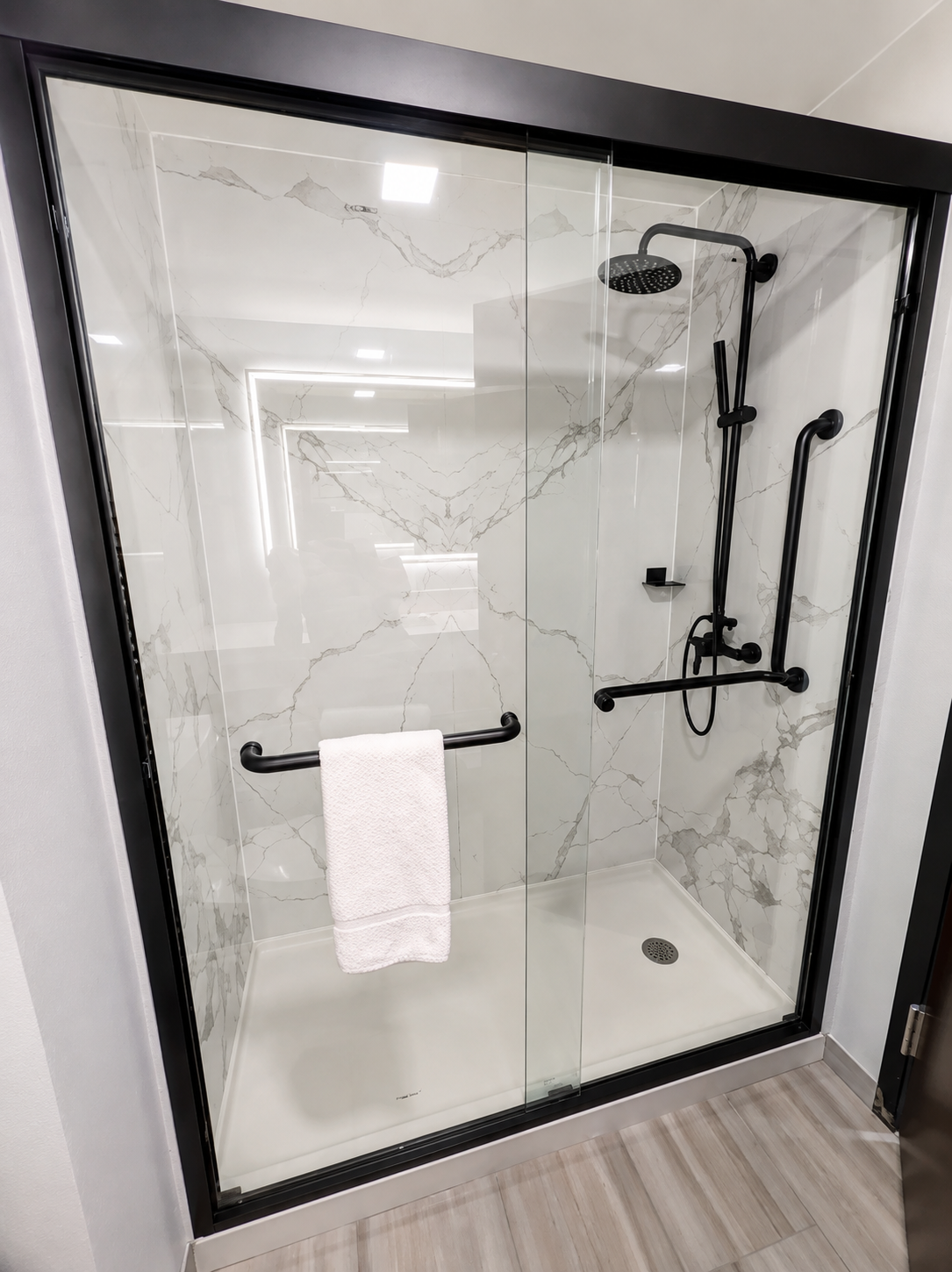 A modern walk-in shower enclosure with a black metal frame and clear glass doors, featuring a white marble-look tiled shower floor and walls with a black handheld shower fixture and black towel bar.